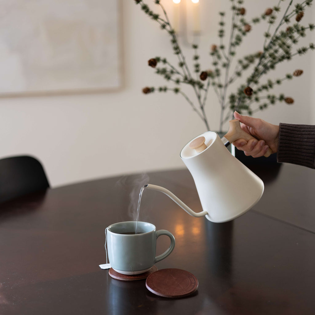 Person pouring steaming hot liquid from a white kettle into a mug that is sitting on a custom logo coaster.
