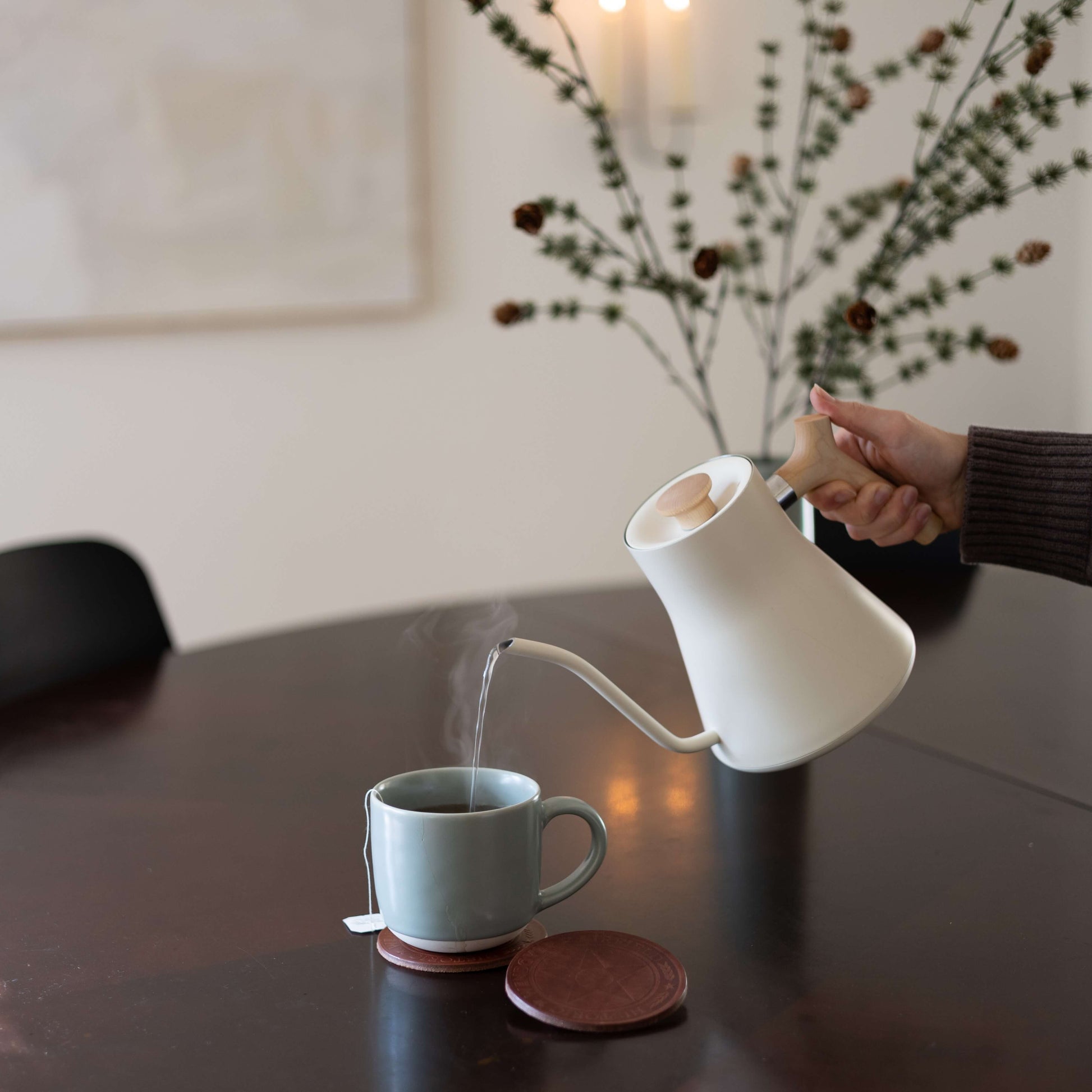 Person pouring steaming hot liquid from a white kettle into a mug that is sitting on a custom logo coaster.