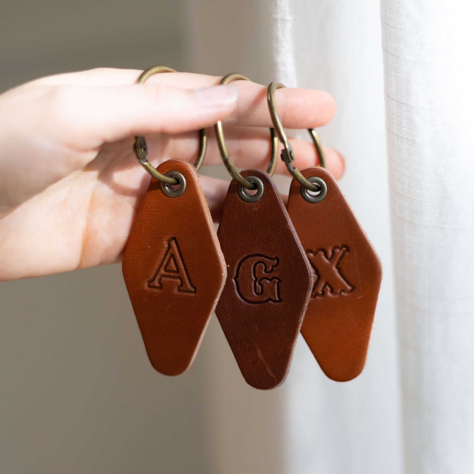 Hand holding three brown leather key tags with custom embossed initials against a neutral background